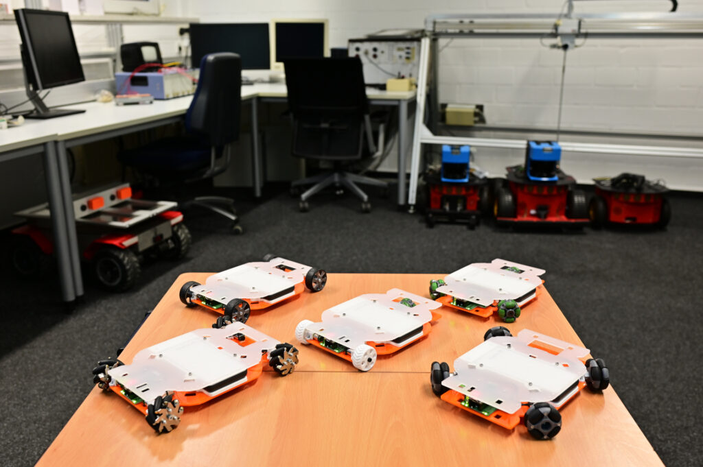 Fünf EduRobs unterschiedlicher Kinematiken (Skid Steering, drei OmniWheels, vier Omniwheels, Mecanum und Differentialantrieb mit Omniwheel-Stützard) auf Tisch angeordnet. Im Hintergrund sind mehrere alte Roboter unscharf abgebildet.