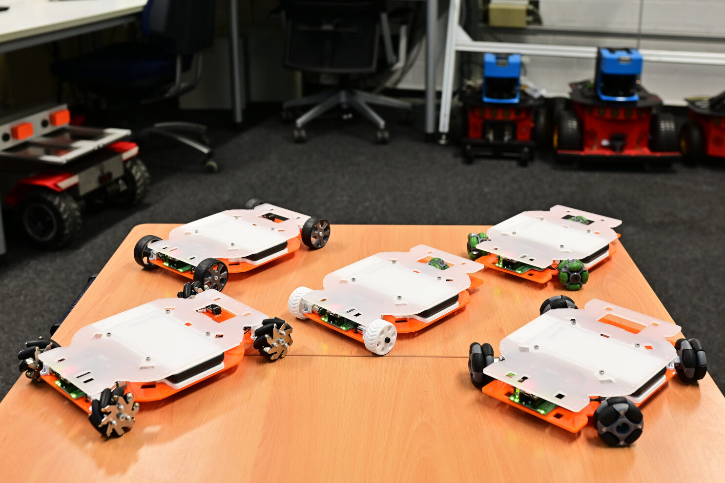 Fünf EduRobs unterschiedlicher Kinematiken (Skid Steering, drei OmniWheels, vier Omniwheels, Mecanum und Differentialantrieb mit Omniwheel-Stützard) auf Tisch angeordnet. Im Hintergrund sind mehrere alte Roboter unscharf abgebildet.